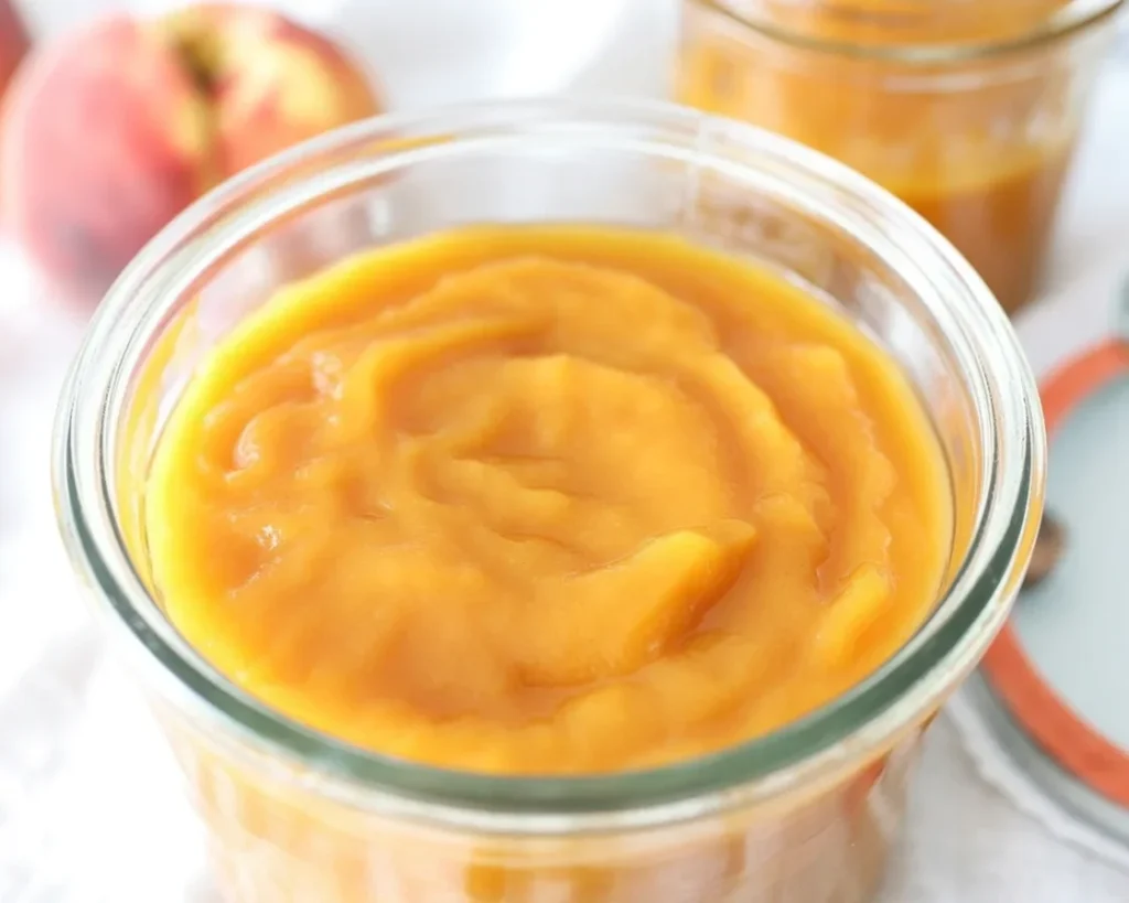 Jar of homemade peach butter with fresh peaches and ingredients beside it.